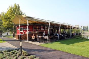 An outdoor restaurant with a beige canopy ober the seating area, a green lawn in front of it