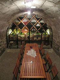  a long cherry stained wooden bench with chairs inside a vaulted stone cellar