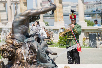 a trumpeter in hussar costume at a bronze statue and fountain