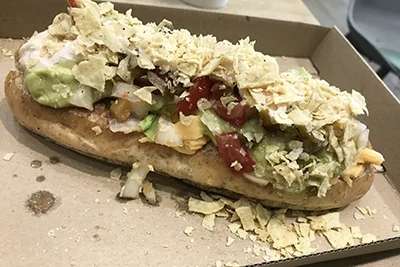 Mexican style vegan hotdog on a cardboard tray - seitan frankfurter on spelt baguette, guacamole, sprinkled with tortilla