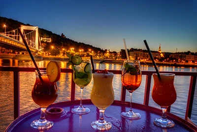 5 different cockatils on a round tabletop on the open deck of a cruise boat at sunset, Elizabeth Bridge can be seen in the background