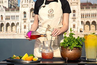 a young female bar mixer mixing a long drink in a glass pitcher on board a boat in daytime, the Parliament can be seen behind her back