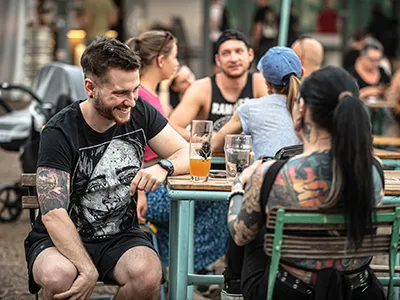 A heavily tattoed man and woman with long black hair sitting at a table with berr glasses in front of them, possibly in a garden of a bar