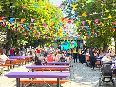 festival atmosphere in City Hall park in May: people sitting at purple benches eating, drinking, talking, there1s a concert on the stage in the background
