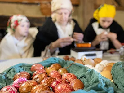 an older woman and two teenage grils dressed in folk costume decorating easter eggs, a a dozen or so painted eggs mainly in red and pruple color on a teal textile in the foreground