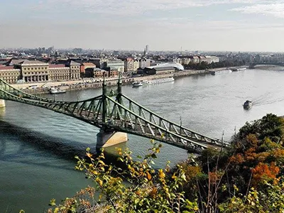 A scenic panoramic view of the Danube River in Budapest probably early autumn. The green Liberty Bridge crosses the foreground, with the city's buildings lining the riverbank in the distance under a hazy autumn sky.