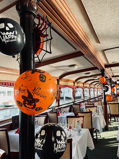 The interior of a dining boat is decorated for Halloween, with black and orange balloons hanging from a support pillar. White tablecloths and table settings are visible in the background. The interior of a dining boat is decorated for Halloween, with black and orange balloons hanging from a support pillar. White tablecloths and table settings are visible in the background.