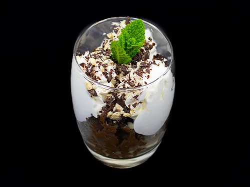 A dessert glass filled with rich avocado chocolate mousse, topped with whipped cream, chocolate shavings, chopped nuts, and a sprig of fresh mint, set against a black background.