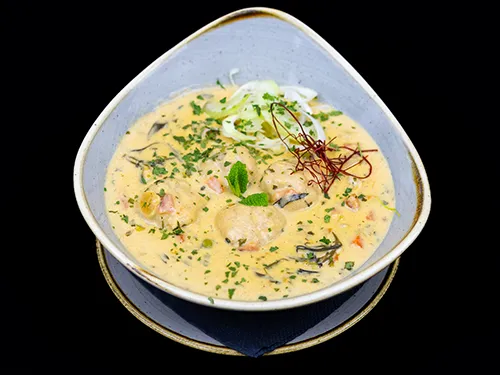 A bowl of creamy tarragon chicken meatball soup garnished with fresh herbs, thinly sliced vegetables, and chili threads, served in a gray ceramic bowl on a dark background.