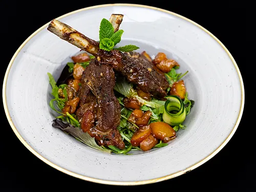 A plated dish of glazed lamb chops served on a bed of mixed greens and sautéed vegetables, garnished with cucumber ribbons and a sprig of fresh mint, presented in a light gray bowl against a dark background.